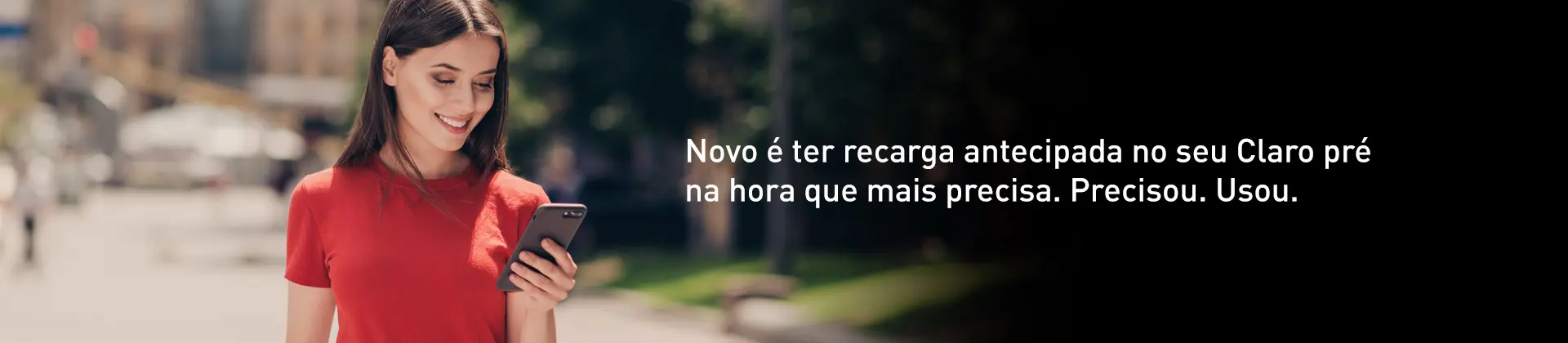 Imagem em ambiente externo de mulher jovem, cabelos lisos, pele clara, camiseta vermelha, sorrindo e olhando para o celular. Texto na imagem: Novo é ter recarga antecipada no seu Claro pré na hora que mais precisa. Precisou. Usou.