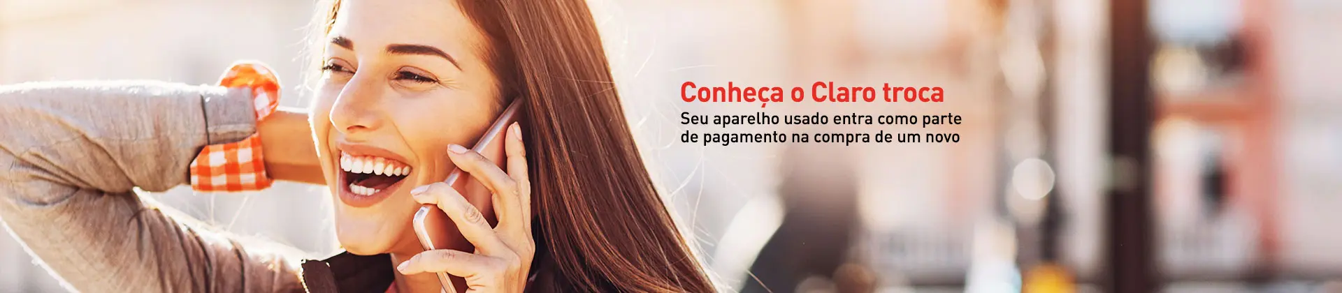 Foto em ambiente externo durante o dia. Mulher jovem com cabelos longos e lisos de cor escura, pele clara, sorrindo e falando ao celular. 