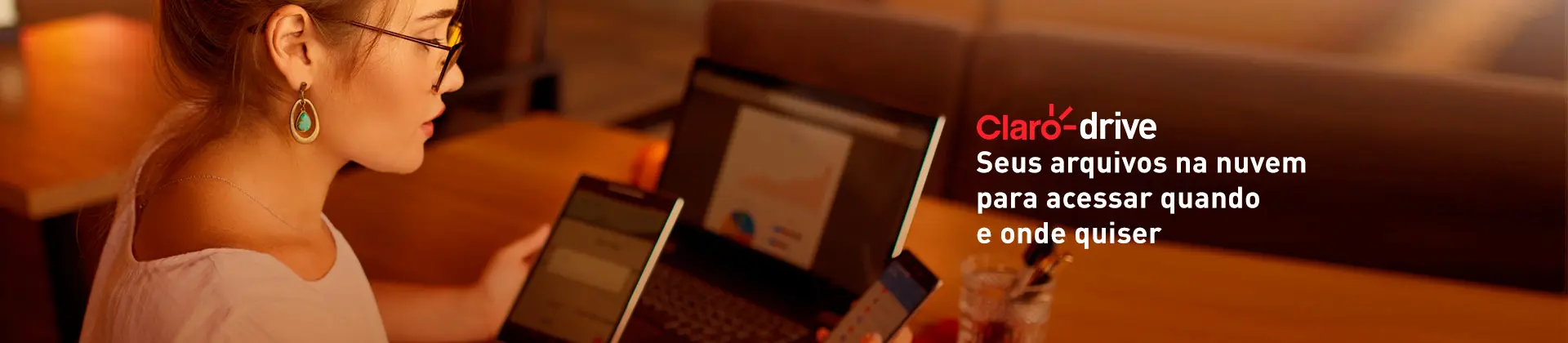 Mulher com tablet e celular nas mãos e notebook aberto sobre a mesa.