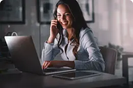 Mulher jovem, morena, sorri, enquanto fala ao telefone. Ela também está usando o notebook à sua frente.