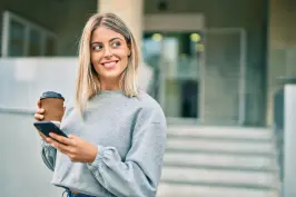 Uma moça de cabelos loiros com um celular na mão sorrindo e olhando para o lado.