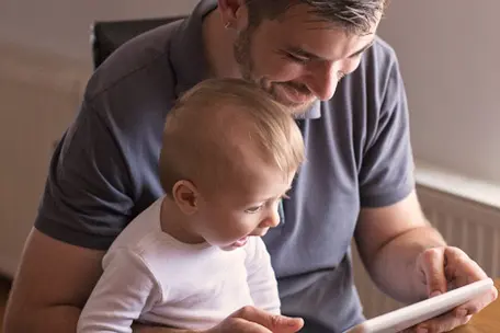 Imagem de um pai com um bebê no colo mexendo em um tablet.