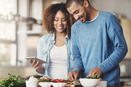 Imagem de um casal cozinhando juntos. a Moça segura um celular na mão enquanto o homem prepara os alimentos.