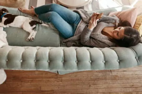 Uma moça e um cachorro deitados no sofá verde enquanto mexe no celular.