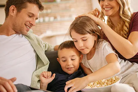 Imagem de uma família com marido, esposa e dois filhos comendo pipoca e mexendo num tablet.