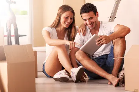 Imagem de um casal sorrindo olhando para um tablet sentados no chão.