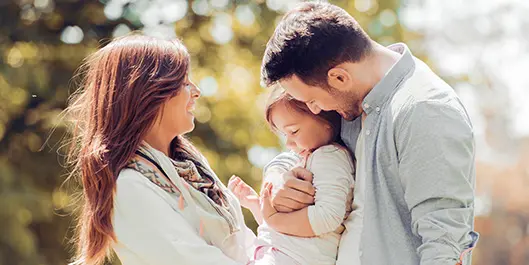Imagem de uma família, a mulher, a filha e o homem. A mulher carrega a menina no colo e o homem a abraça.