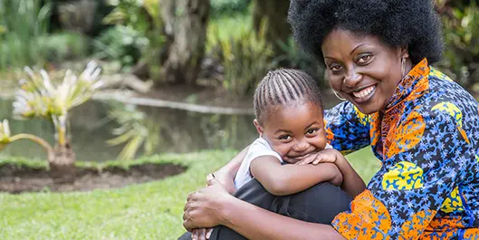 Imagem de mãe e filha negras sorrindo com árvores e um lago no fundo