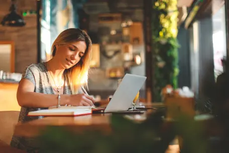 Imagem de mulher escrevendo em um caderno em frente a um notebook.