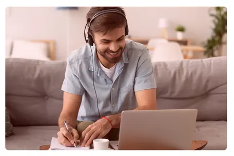 Homem alegremente com fone de ouvido e fazendo anotações em um caderno enquanto observa um notebook.