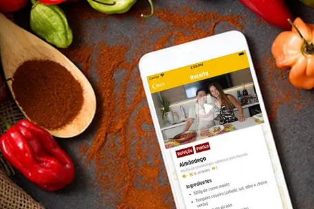 Imagem de um celular com uma receita do Você Gourmet na tela. O celular está sob uma mesa com outros alimentos e temperos em volta.